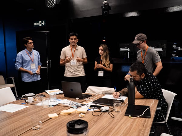 A group of five people are engaged in a collaborative workshop or meeting around a wooden table cluttered with laptops and notes.