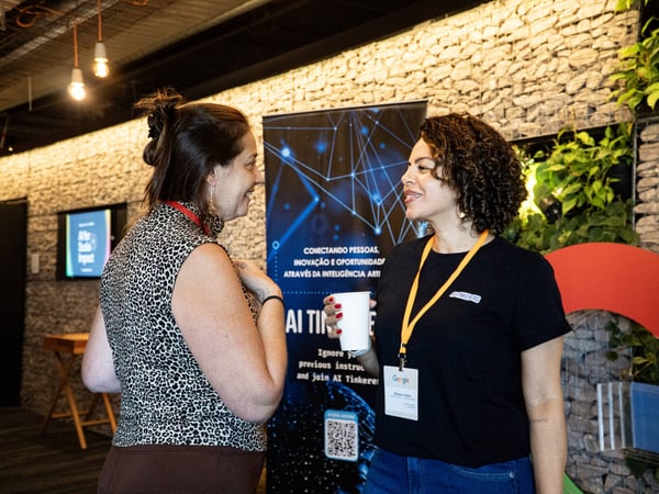 Two women are engaged in a conversation during a networking event, standing in front of a stone wall and a promotional banner for AI Tinkerers.