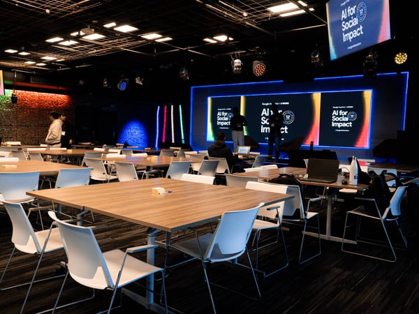 A wide shot of an indoor event space prepared for a hackathon, featuring numerous wooden tables, white chairs, and a stage with large digital screens.