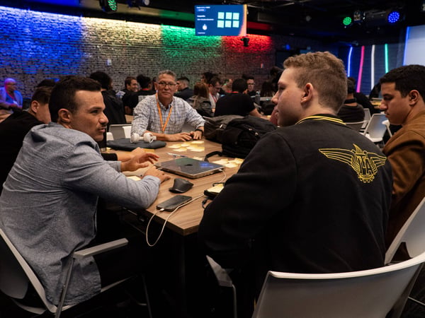 A group of people are gathered around tables in a dimly lit room with a brick wall illuminated by colorful lights, seemingly engaged in a workshop or discussion.