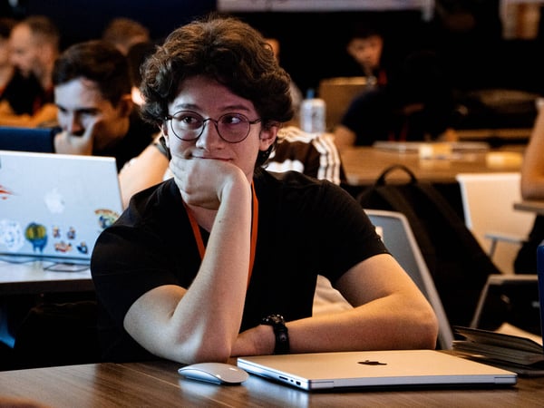 A young man with curly hair and glasses sits at a desk, resting his chin on his hand with a thoughtful expression in a room filled with people and laptops.