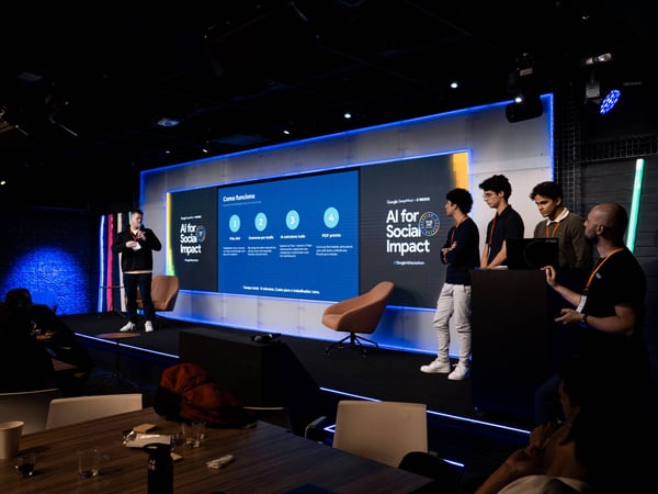 A group of people are on a stage during a presentation for 'AI for Social Impact' at a Google AI Hackathon. A large screen displays a process flow in Portuguese about transforming messages into resumes using AI.