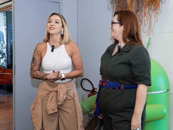 Two women are standing and talking in an indoor setting, with one wearing a white tank top and tan pants and the other in a dark green jumpsuit.