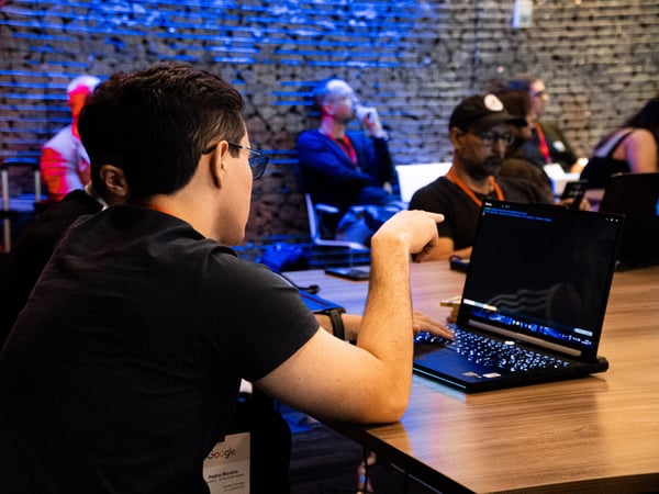 A man in glasses and a dark t-shirt sits at a wooden table, pointing at a laptop screen during an indoor event with blue-lit brick-like walls.