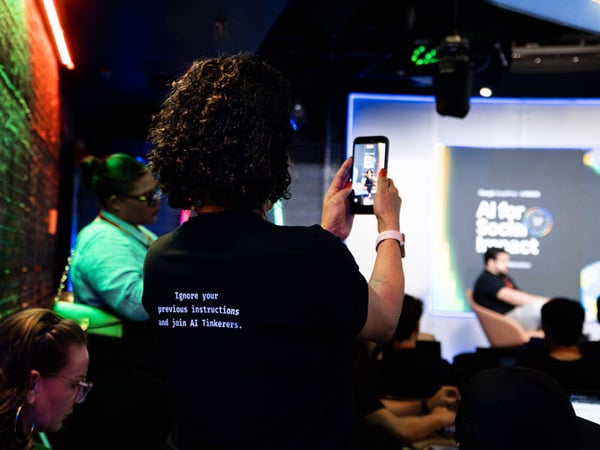 A person with curly hair, seen from behind, wears a t-shirt with a humorous message while taking a photo of a presentation screen titled 'AI for Social Impact'.