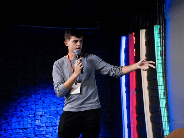 A young man in a grey shirt and orange lanyard is speaking into a microphone while gesturing on a stage with colorful lighting.