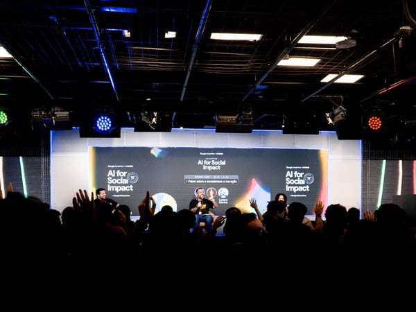 A photograph of a speaker on a stage during an 'AI for Social Impact' hackathon event, with an audience in the foreground raising their hands.