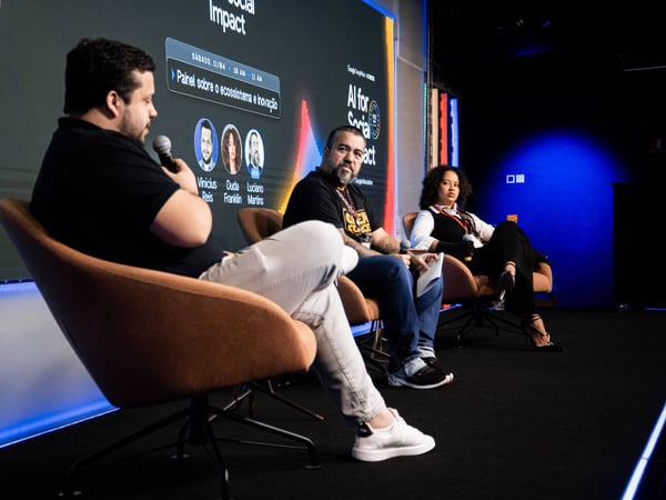 A panel discussion at an event featuring three speakers sitting on a stage in front of a large screen displaying event details.