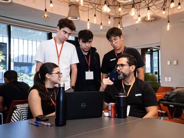A group of young professionals or students are gathered around a laptop in a modern indoor setting, with one man gesturing as if explaining something to the others.