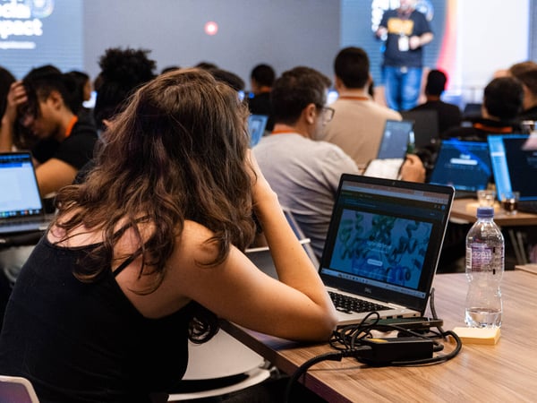 A photograph showing an audience of people with laptops attending a presentation, with a speaker visible on a stage in the background.