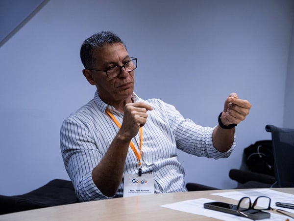 A man wearing glasses and a striped shirt sits at a table, gesturing with his hands as if explaining a concept during a discussion.