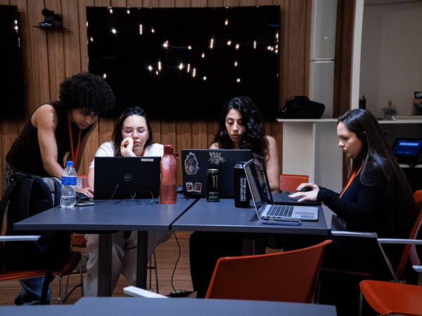 Four women are focused on their laptops while working together at a table in a modern indoor space with wood-paneled walls.