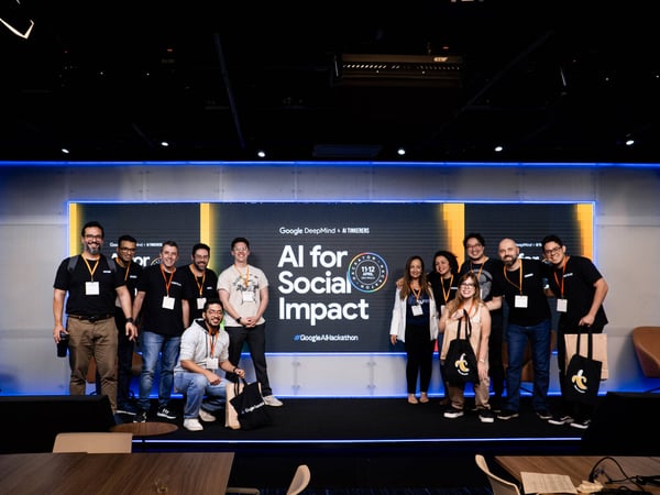 A group of twelve people posing for a photo on a stage in front of a large digital screen at a Google AI Hackathon event.