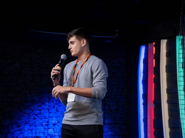 A young man is speaking into a microphone while standing on a stage with a dark textured wall and colorful accent lighting.
