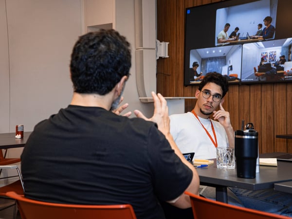 Two men are engaged in a discussion in an office setting, with one man gesturing while the other listens. A large screen in the background displays a multi-person video conference call.