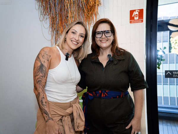 Two women are standing together and smiling for a photo indoors, both wearing lapel microphones.