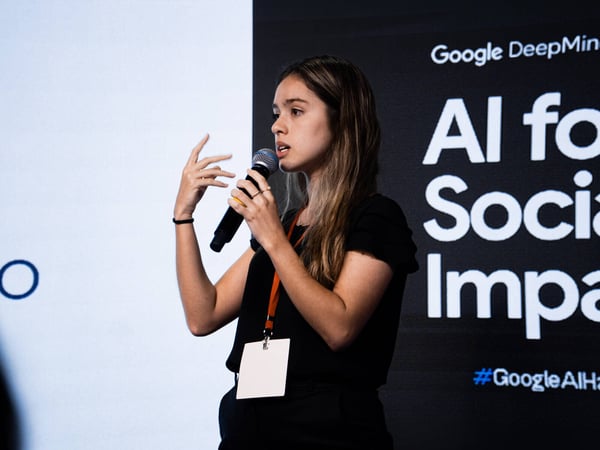 A young woman is speaking into a microphone during a presentation, standing in front of a black backdrop with Google DeepMind branding.