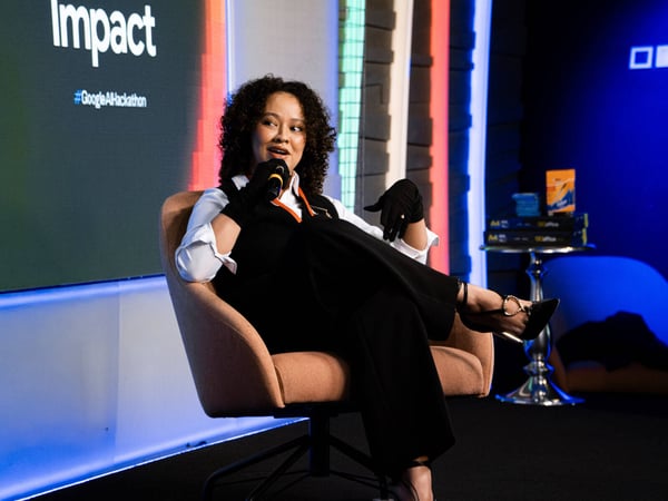 A woman with curly hair sits in a beige armchair on a stage, speaking into a microphone during an event.