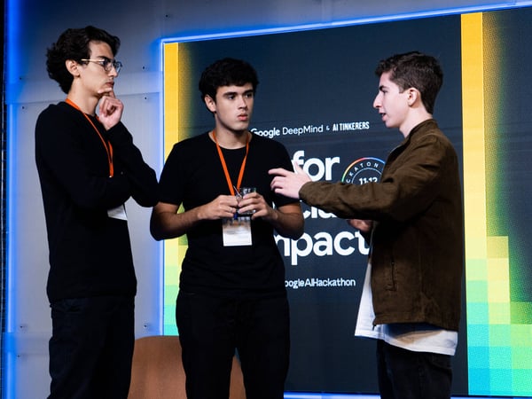 Three young men are engaged in a conversation in front of a digital screen at a hackathon event.