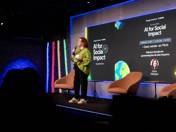 A woman in a green shirt is giving a presentation on a stage, standing in front of a large screen displaying information about an 'AI for Social Impact' hackathon.