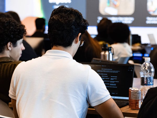 A young man in a light blue polo shirt sits at a desk using a laptop during a presentation or workshop, with other attendees visible in the background.