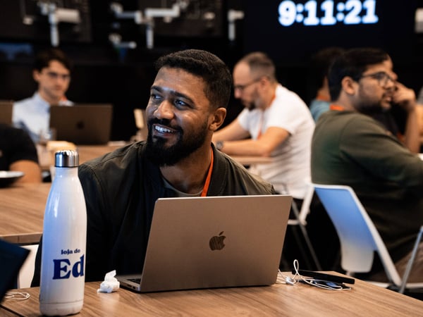 A man with a beard smiles while sitting at a wooden table with a laptop in a busy indoor workspace.