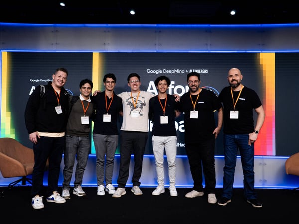 A group of seven men posing for a photo on a stage in front of a digital screen that reads 'AI for Impact'.