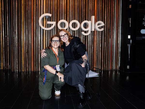 Two women are posing for a photograph in front of a large, white 3D Google logo mounted on a wall of vertical metallic rods.