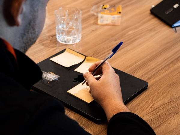 A person is writing on a yellow sticky note placed on top of a closed black laptop on a wooden table.