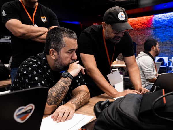 A candid photograph showing several men focused on a laptop screen during a tech event or hackathon in a dimly lit indoor setting.