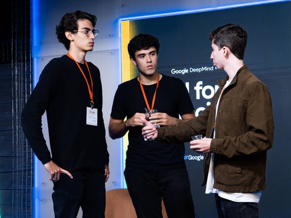 Three young men are engaged in a conversation at a Google DeepMind event, with one man on the right gesturing with a thumbs-up.