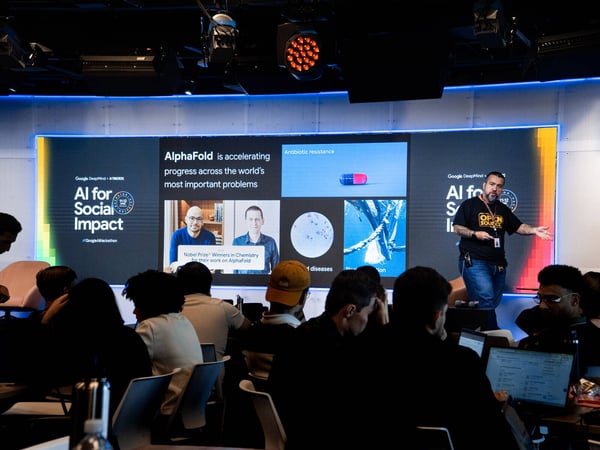 A man stands on a stage giving a presentation about AI for Social Impact, with a large screen behind him displaying information about AlphaFold.