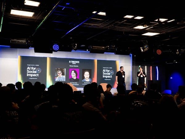 A live presentation event titled 'AI for Social Impact' by AI Tinkerers in São Paulo, showing speakers on stage and an audience in a dark auditorium.