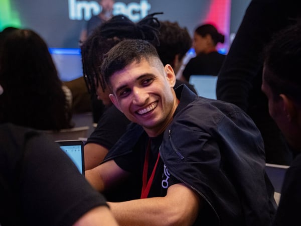 A young man with a bright smile is sitting at a table during an indoor event or workshop, surrounded by other participants and laptops.