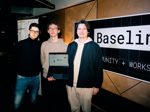 Three young men pose together indoors, with one holding an open laptop, standing in front of a sign that reads 'Baseline COMMUNITY + WORKSPACE'.