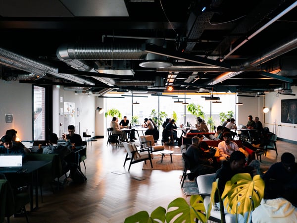 A wide-angle photograph of a spacious, modern co-working space filled with people working on laptops, chatting, and relaxing. The room features industrial elements like exposed ceiling pipes, warm wooden floors, and large windows that let in natural light.