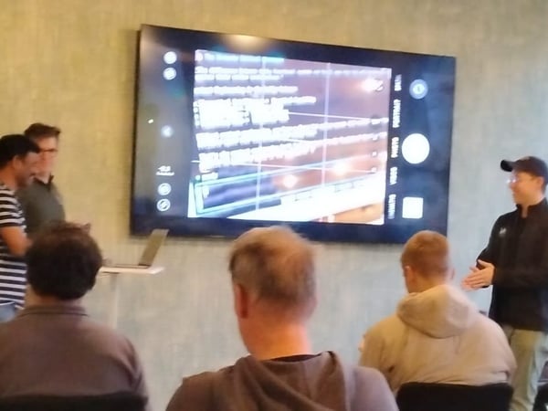 A candid photograph taken indoors showing a technical presentation where a group of people is gathered around a large display screen that is showing a mobile camera interface or similar application.