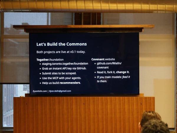 A photograph showing a digital screen displaying a presentation slide titled 'Let's Build the Commons' in an indoor setting with brick walls.