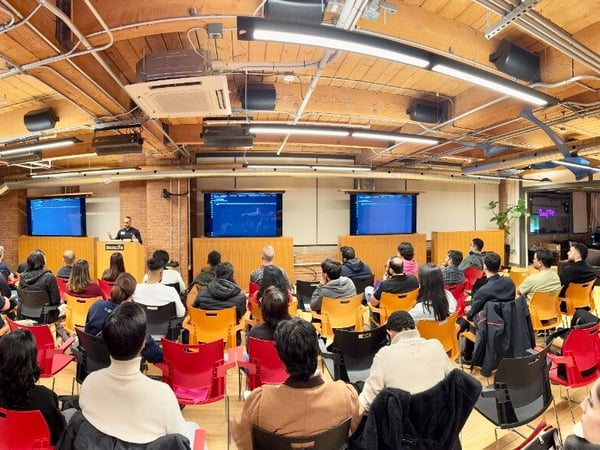 A wide-angle photograph of a tech presentation taking place in a modern office space with an exposed wooden ceiling, featuring a speaker at a podium and a large seated audience.