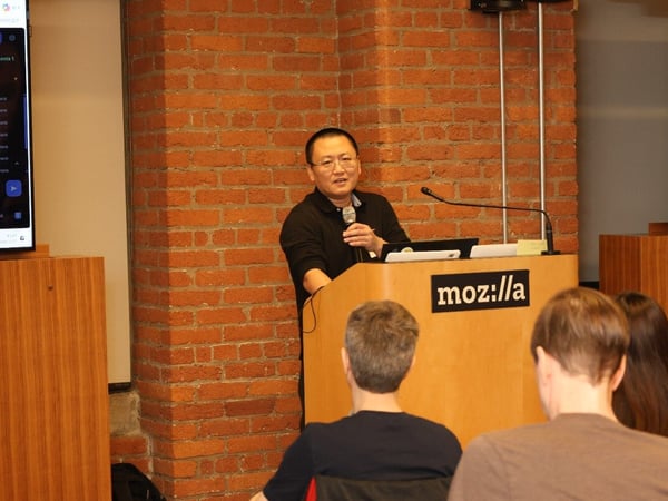 A man is giving a presentation behind a wooden podium with the Mozilla logo in a room with a brick wall.