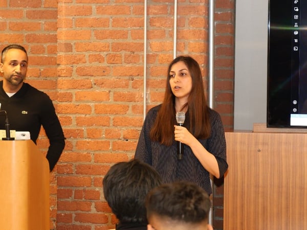 A woman is speaking into a microphone during a presentation in an indoor setting with a brick wall background, while an audience listens.