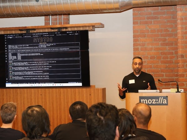 A man stands at a podium with a Mozilla logo, giving a tech presentation in front of an audience, with a large screen displaying code behind him.