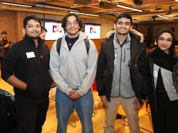 A group of four young adults, three men and one woman, are standing and smiling together in an indoor event space with wooden floors and exposed ceiling pipes.