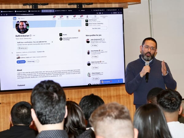A man stands at the front of a room, speaking into a microphone while gesturing. Behind him, a large screen displays the LinkedIn profile of Raffi Krikorian, while an audience sits in the foreground.
