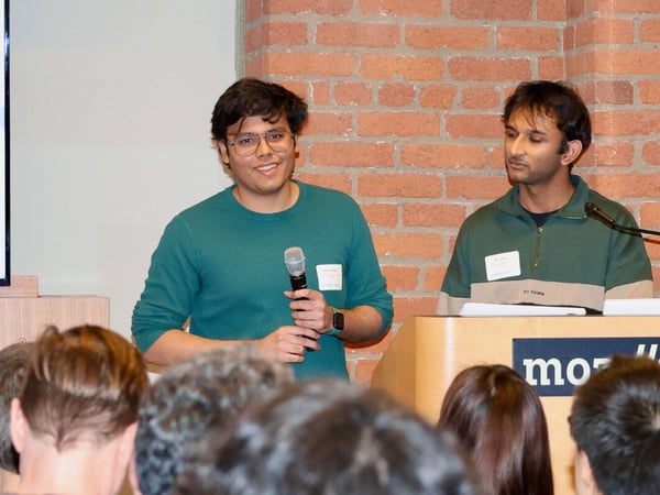 A man in a teal sweater is speaking into a microphone during a presentation, standing next to another man at a podium against a brick wall background.