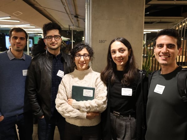 A group photo of five young professionals, three men and two women, smiling directly at the camera while standing indoors at what appears to be a networking or corporate event.