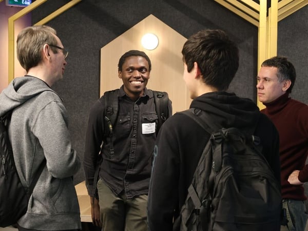 Four men are gathered indoors, appearing to be engaged in a casual conversation in a modern venue featuring gray acoustic panels and yellow wooden architectural elements. The man in the center is smiling brightly and wearing a name tag.
