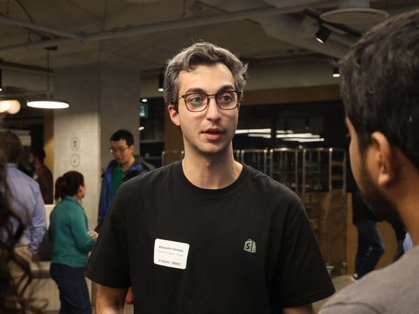 A candid photograph shows a man wearing glasses and a black Shopify t-shirt, identified by his name tag as Alexandre Garepy, speaking to another individual at an indoor professional gathering.