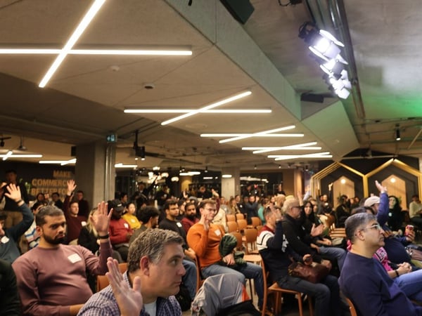 A candid photograph captures a large audience seated in an indoor event space, with dozens of attendees raising their hands, likely responding to a question or participating in a poll.