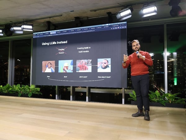 A man wearing an orange sweater and dark pants is giving a presentation on a stage, holding a microphone and gesturing toward a large screen behind him. The screen displays a dark slide outlining a process related to LLMs, and the event takes place indoors with large windows revealing a nighttime city view.